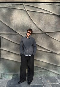 Jeune homme portant des lunettes de soleil, une veste grise zippée et un pantalon large noir, debout les mains dans les poches devant un mur en pierre texturé aux formes géométriques.