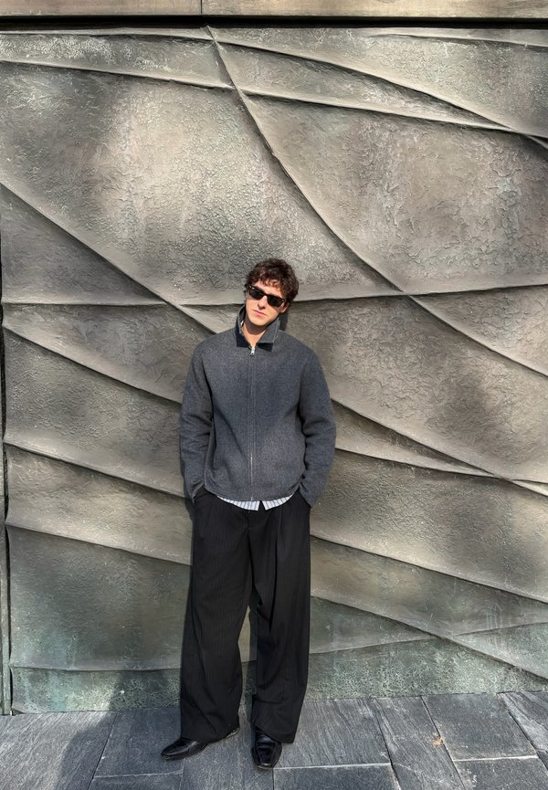 Fiatal férfi napszemüvegben, szürke cipzáras dzsekiben és fekete, bő nadrágban, kezét a zsebében tartva áll egy texturált, geometrikus kőfallal szemben.