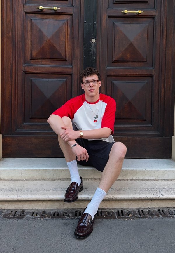 Rojo y blanco camiseta de raglán con un gráfico de cereza, pantalones cortos negros, calcetines blancos y mocasines burdeos; sentado en los escalones cerca de puertas de madera.