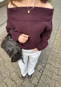 Žena u ljubičastom puloveru s otvorenim ramenima, bijelim hlačama, smeđim cipelama, zlatnom ogrlicom u obliku srca, drži crnu kožnu torbu na pločniku od kamenih kocki.