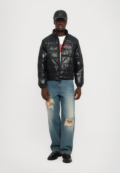 Chaqueta de plumas negra con textura brillante, cierre de cremallera y logo rojo. Lleva puesta sobre una camiseta blanca y se combina con jeans azules desgastados y zapatos negros.