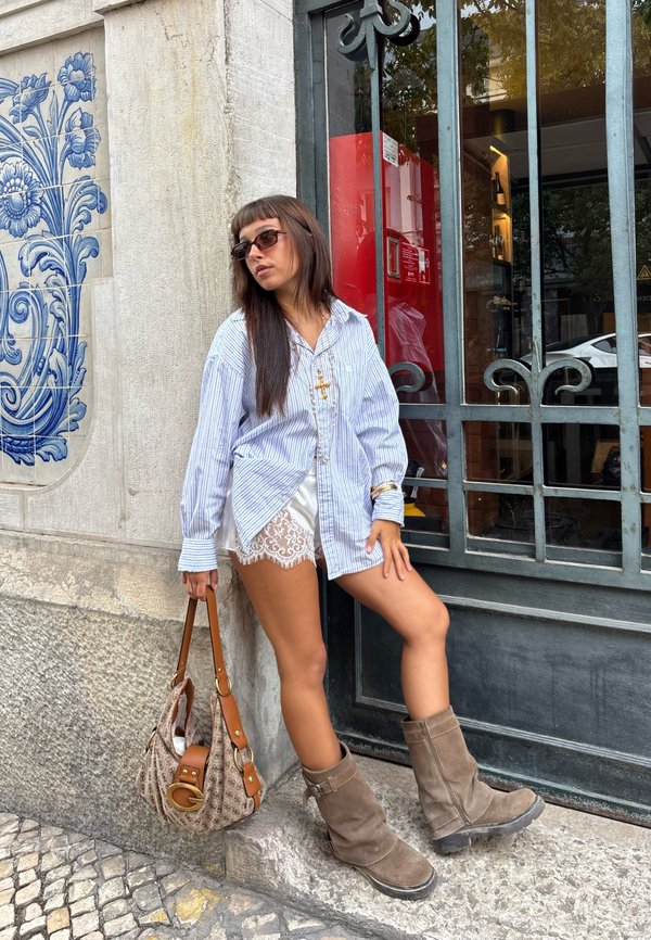 Femme portant des lunettes de soleil, une chemise rayée surdimensionnée, des shorts en dentelle et des bottes marron, appuyée contre un mur avec des carreaux bleus, tenant un sac à main à motifs.