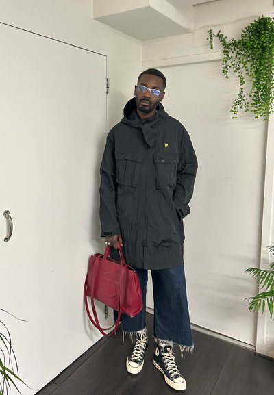 Hombre con chaqueta negra, vaqueros anchos y cortos, zapatillas negras de caña alta, sosteniendo un bolso rojo, de pie en el interior cerca de una puerta blanca y plantas.