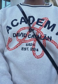 Maglione grigio a maglia con il testo in rosso e blu "ACADEMY" e una grafica di una corda. La tracolla di una borsa è visibile sul davanti.