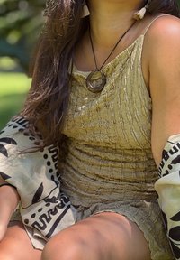 Femme aux longs cheveux portant une robe beige texturée, un collier pendentif en bois, des boucles d'oreilles à pampilles et un châle à motifs en plein air sous la lumière naturelle.