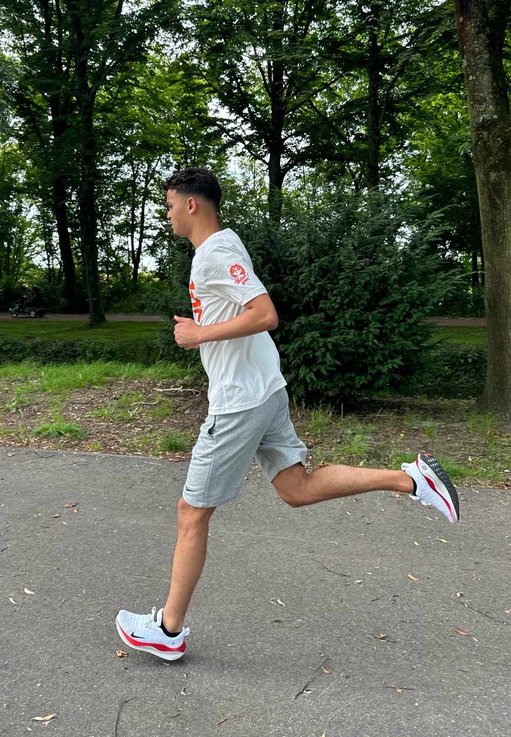 T-shirt de sport blanc avec logo rouge, shorts gris et chaussures de course blanches avec accents rouges, capturé en pleine foulée sur un chemin du parc.
