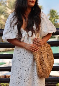Round, woven straw bag with a textured surface, held by a person wearing a white dress with eyelet patterns and puff sleeves.