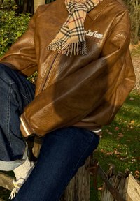 Brun læder bomberjakke med ribbede manchetter og krave. Kombineret med mørkeblå jeans og et beige ternet tørklæde med frynser.