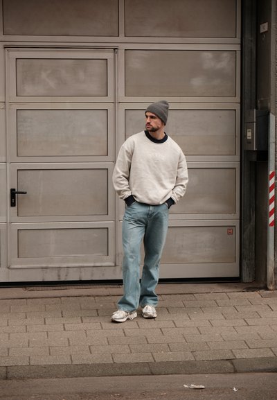Sudadera gris clara con cuello redondo, detalles en azul y logo, combinada con unos vaqueros sueltos de color azul claro y zapatillas grises, frente a una puerta de garaje.