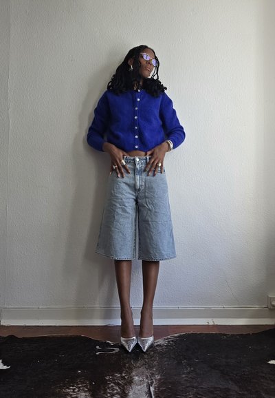 Cardigan azul de terciopelo, culottes de mezclilla de tiro alto y zapatos de tacón plateados. Los accesorios incluyen anillos y gafas de sol. Fondo de pared lisa.