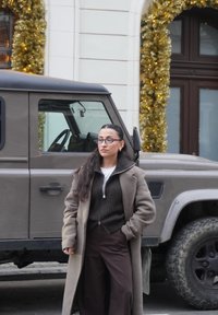 Femme portant un manteau marron sur un pull foncé et un pantalon large, debout à côté d'un SUV sombre à finition texturée. Feuillage décoratif doré en arrière-plan.