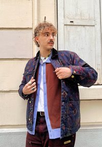 Ναυτική patterned ζακέτα με λουλουδάτα σχέδια, καφέ υφασμάτινο πουκάμισο από κάτω, σε στρώσεις πάνω από ένα ανοιχτό μπλε πουκάμισο με κουμπιά, φορεμένο με σκούρες κόκκινες παντελόνες.