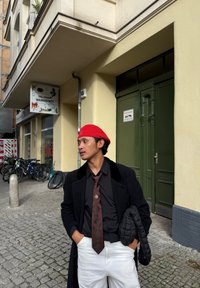 Cappotto nero con colletto in velluto, camicia nera, cravatta marrone a motivi, pantaloni bianchi e berretto rosso. Sullo sfondo una porta verde e delle biciclette.