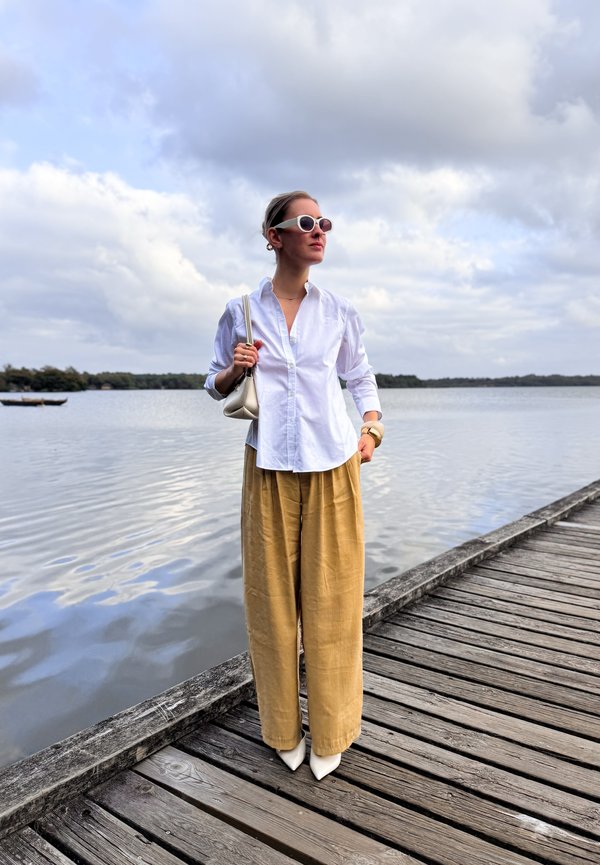 Mujer con blusa blanca, pantalones anchos beige y gafas de sol, de pie en un muelle de madera junto a un lago tranquilo bajo un cielo nublado.