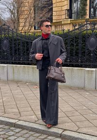 Homme en costume gris foncé et foulard rouge autour du cou tenant un sac à main en cuir texturé foncé, debout sur un trottoir devant une clôture en métal noir ornée.