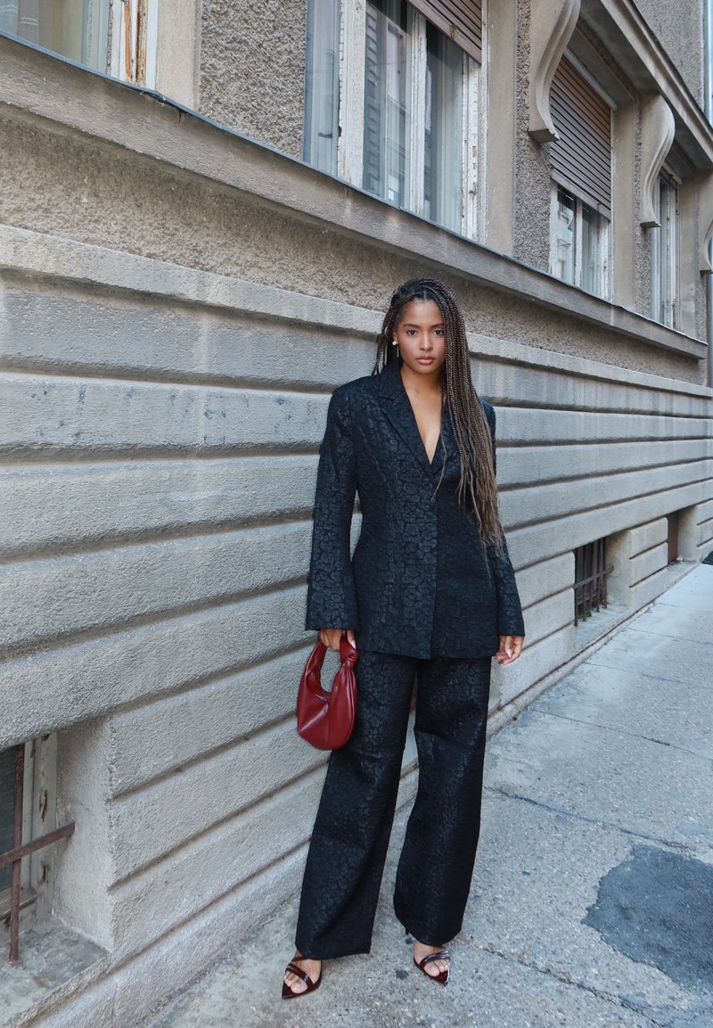 Svart texturerad kostym med vida byxor och en skräddarsydd kavaj. Accessoarer inkluderar en röd handväska och svarta remsandaler med klack. Betongbakgrund.