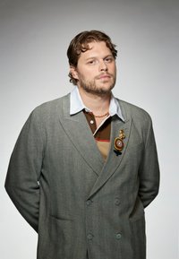 Grå dobbeltbrystet blazer med et strukturstof, båret over en brun og stribet skjorte. Har en dekorativ brosche på klappen.