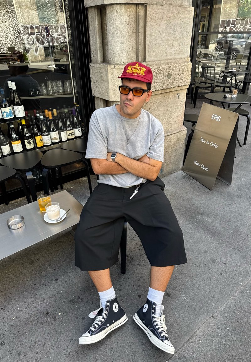Personne portant une casquette bordeaux, une chemise grise et un short noir jusqu'aux genoux, assise à côté d'une table avec des boissons. Baskets montantes noires et chaussettes blanches.