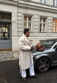 Kremfarget frakk, hvite bukser, brune lærhansker, brun veske på en grå bil med røde bremsekalipere, foran en bygning.