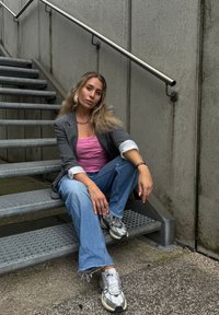 Szary żakiet na różowej, teksturowanej bluzce, niebieskie spodnie dzwony i srebrne sneakersy. Metalowa klatka schodowa i betonowa ściana w tle.