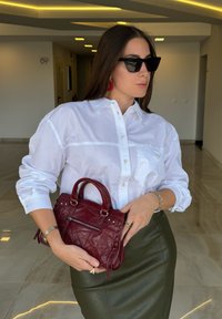 White button-up shirt, green leather skirt, red accessories, holding a small burgundy leather handbag with silver hardware and a zip pocket.