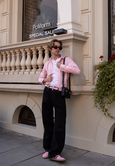 Camisa de botón rosa claro sobre una camiseta blanca tipo henley, combinada con vaqueros anchos negros y zapatillas rosas. Bolsa mensajera negra y gafas de sol.