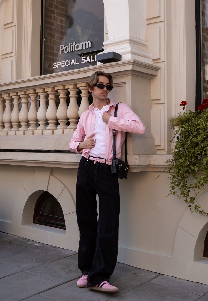 Camisa de botão rosa claro sobre uma henley branca, combinada com jeans largas pretas e ténis cor de rosa. Bolsa de mensageiro preta e óculos de sol.