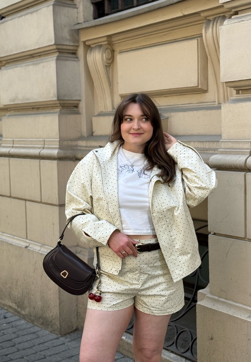 Jeune femme portant une veste légère à pois et un short, un crop top blanc, tenant un sac à main noir, debout près d’un mur beige d’un bâtiment.