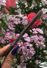 Hand die een bruine NYX eyelinerpotlood vasthoudt boven roze en witte bloemen met rode rozen op de achtergrond.