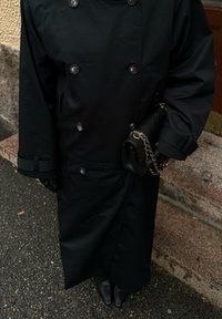 Black double-breasted coat with shiny buttons, side pockets, and cuffs. Paired with a black quilted handbag featuring a gold chain.