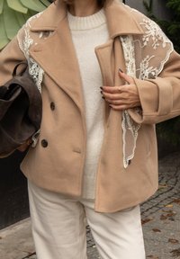 Béžový dvojradový vlnený kabát s čipkovou vrstvou na ramenách, nosený cez ľahký pletený sveter a kombinovaný s krémovými nohavicami.