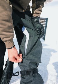 Pantalon de neige noir avec une poche zippée et une sangle ajustable. Une doublure en filet blanche est visible à l'intérieur de la partie non zippée. Arrière-plan enneigé.