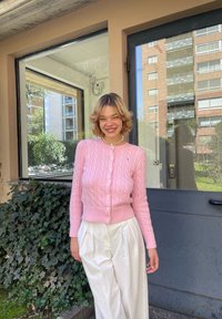 Ανοιχτό ροζ καμπαρντίνα με πλέξη καλωδίων, κουμπωμένη και με ραμμένο στρίφωμα, φορεμένη πάνω από λευκά πλισέ παντελόνια. Το φόντο περιλαμβάνει πράσινο φυλλωσιές και σύγχρονη αρχιτεκτονική.