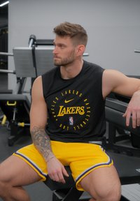 Zwarte mouwloze shirt met 'Lakers' logo, gele shorts met paarse rand, fitte mannelijke model zittend op een gymbank, gymapparatuur op de achtergrond.