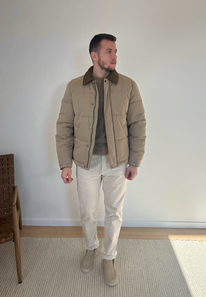 Homme portant une doudoune beige, un pull marron, un pantalon clair et des bottes beiges, debout à l'intérieur sur un tapis à côté d'une chaise en bois.