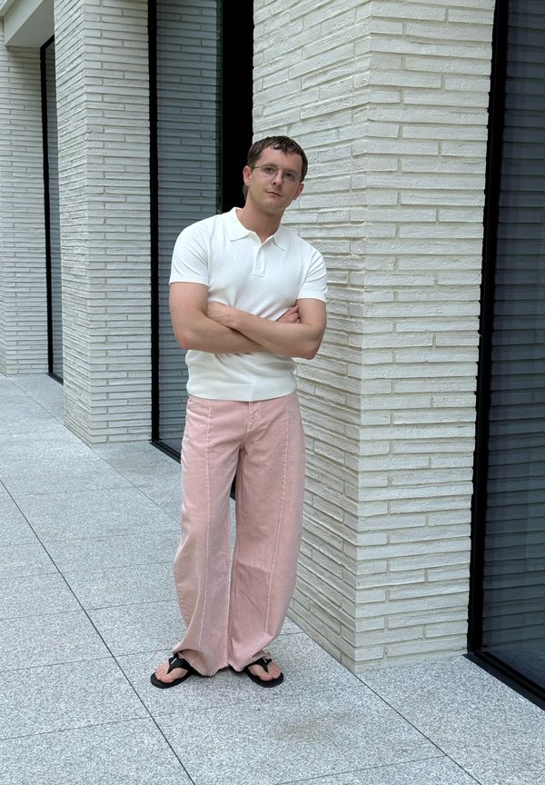 Hombre con gafas está de pie con los brazos cruzados, vistiendo un polo blanco de manga corta, pantalones anchos rosas y sandalias negras junto a una pared de ladrillo blanca.