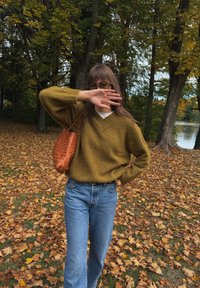 Olive rohelise värvusega kudum, millel on V-kaelus, sinised sirged teksad ja tekstureeritud pruuni kudumisega käekott, ümbritsetud langenud lehtedest.