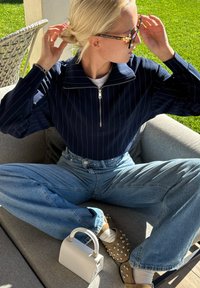 Blondýnka v brýlích si upravuje vlasy, má na sobě námořnicky modrou pruhovanou zipovou mikinu, džíny a hnědé mokasíny se cvočky, zatímco sedí s překříženýma nohama venku.