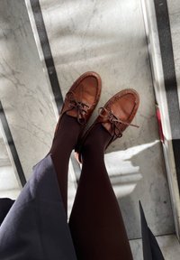Scarpe in pelle marrone con lacci, caratterizzate da una punta arrotondata e dettagli cuciti, indossate con collant scuri su un pavimento di marmo.