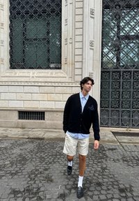 Cardigan noir, chemise bleu clair, shorts crème et mocassins noirs. Debout sur des pavés, avec un bâtiment orné et une fenêtre décorée en arrière-plan.