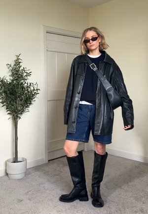 Mujer con chaqueta de cuero negra, pantalones cortos azul marino, botas negras hasta la rodilla, gafas de sol y bolso cruzado, de pie en interiores cerca de una planta en maceta.