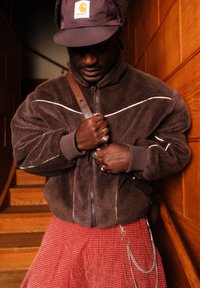 Άνδρας που φορά καφέ φλις μπουφάν, μπορντό καπέλο Carhartt, κόκκινη καρό φούστα, κρατώντας λουρί δερμάτινης τσάντας σε εσωτερικό χώρο δίπλα σε ξύλινη σκάλα.