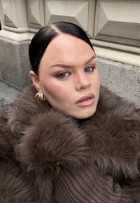 En person med ljus hud och blå ögon bär en tjock brun fuskpälsrock. Rocken har en mjuk, voluminös textur och under bärs en brun ribbad stickad tröja.