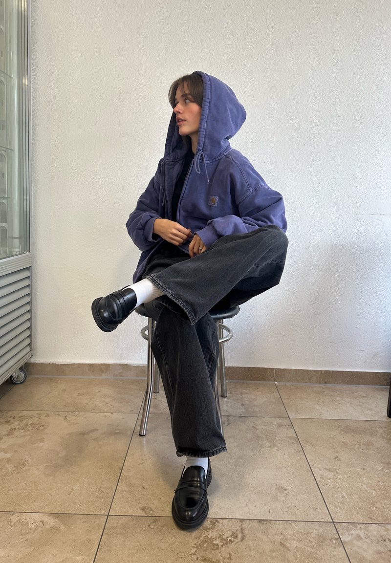 Felpa con cappuccio viola, jeans neri a gamba larga e scarpe nere con calzini bianchi. Modella seduta su uno sgabello, con piastrelle del pavimento visibili.