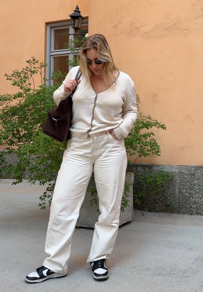 Cárdigan de punto beige con cierre de botones, combinado con pantalones de pierna ancha color crema claro y zapatillas negras. Bolsa marrón en el hombro.