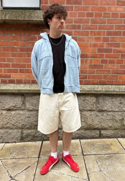 Sudadera con cremallera azul claro, camiseta negra, pantalones cortos blancos y zapatillas deportivas rojas. Fondo de pared de ladrillo y pavimento de concreto.