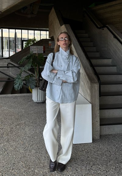 Mujer con chaqueta gris claro, pantalones blancos y gafas, de pie en el interior cerca de unas escaleras de hormigón con los brazos cruzados.