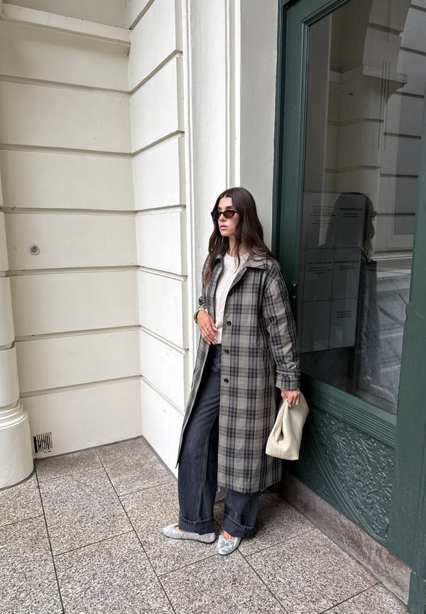 Mujer que lleva un abrigo largo de cuadros gris, pantalones de pierna ancha, gafas de sol y zapatillas plateadas, sosteniendo un bolso blanco, de pie cerca de una puerta verde y una pared blanca.