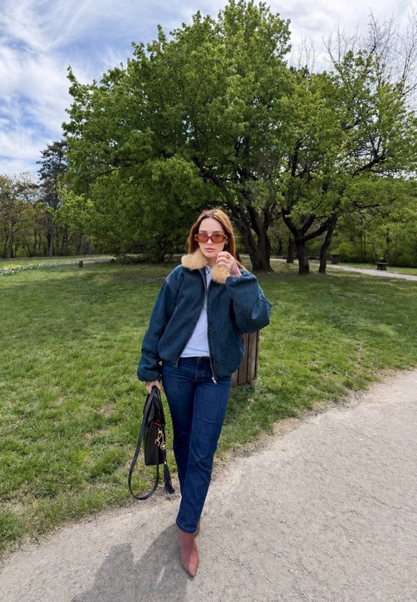 Femme portant des lunettes de soleil, une veste à col en fourrure, un jean et des bottes, marchant sur un chemin de parc tenant un sac à main noir, avec des arbres verts derrière.