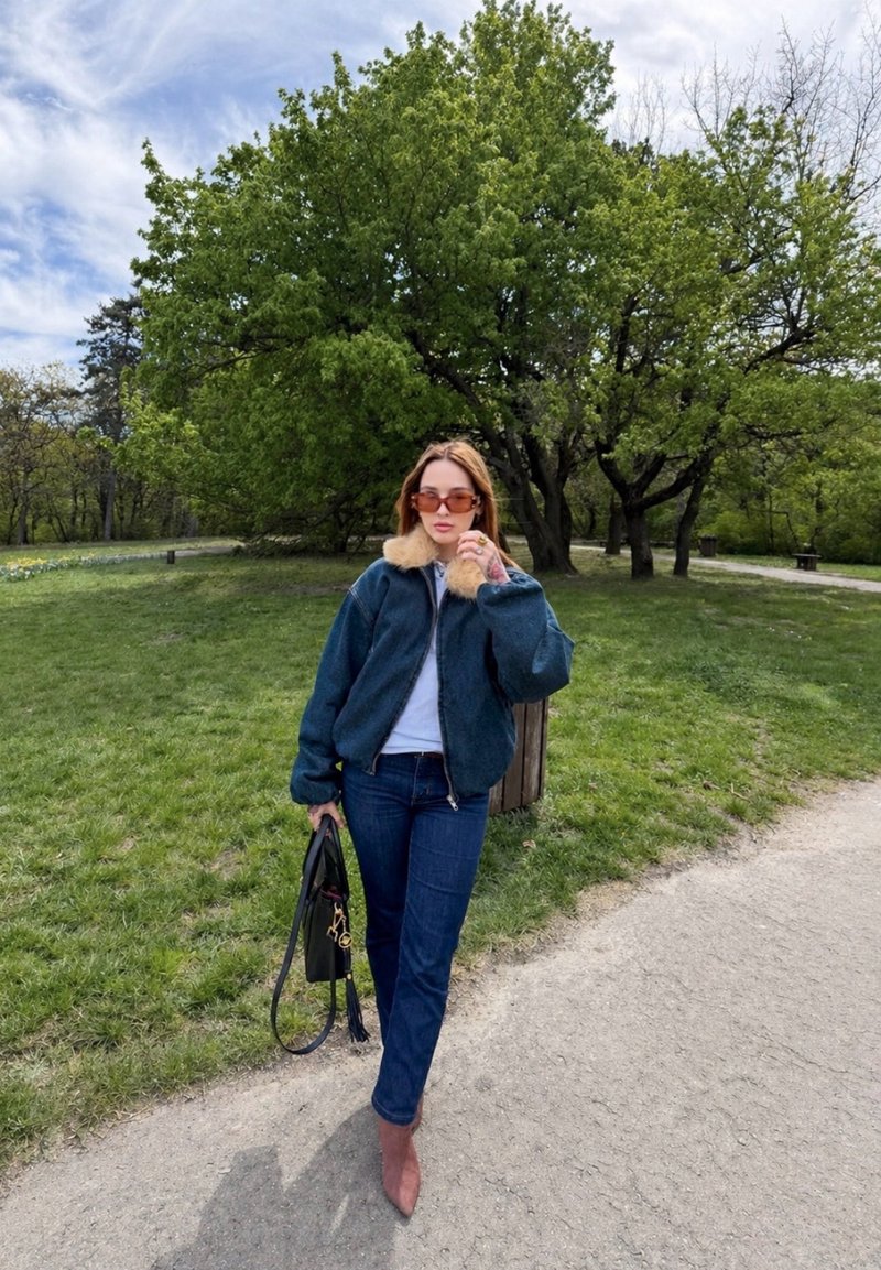 Frau mit Sonnenbrille, Jacke mit Pelzkragen, Jeans und Stiefeln geht auf einem Parkweg und hält eine schwarze Handtasche, mit grünen Bäumen im Hintergrund.